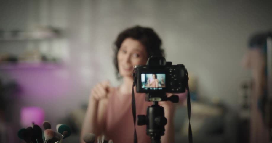 White woman sitting at a table full of makeup brushes and makeup tools talking to a camera recording her