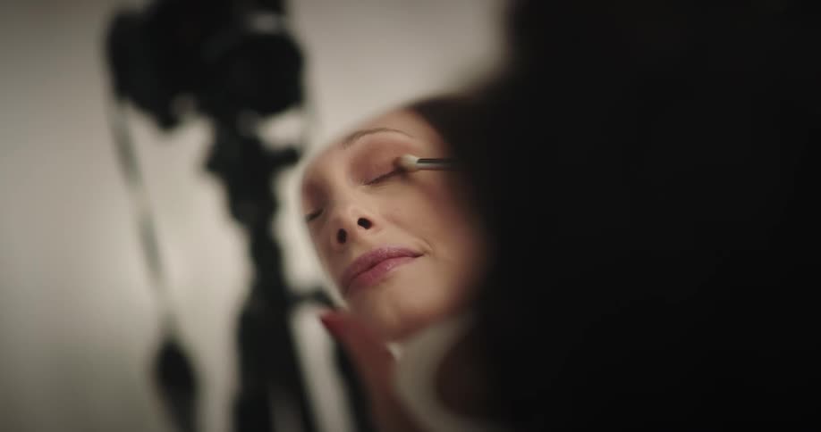 A woman uses a brush to apply eye shadow while looking at her face in a handheld mirror