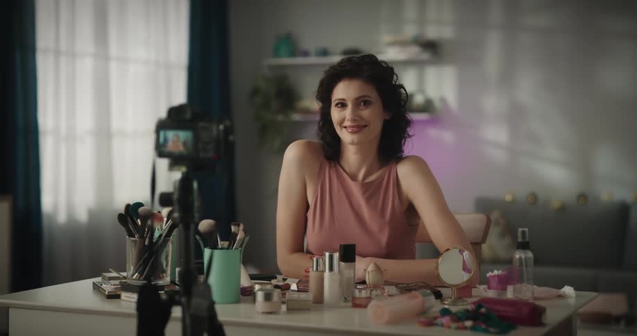 White woman sitting at a table full of makeups and cosmetic products with a digital camera pointed to her