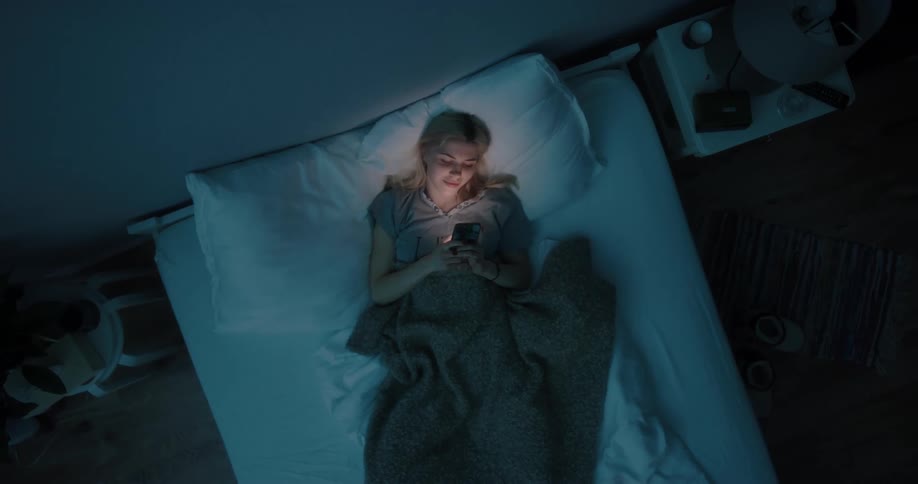 Blonde woman lying down in bed uses smartphone at night in bedroom