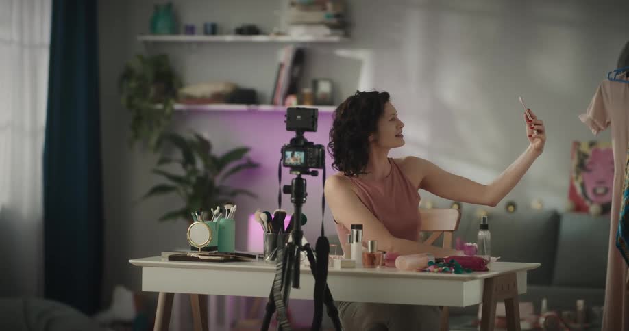 A woman sitting at a table full of cosmetics waves at her phone while filming herself with a camera on a tripod