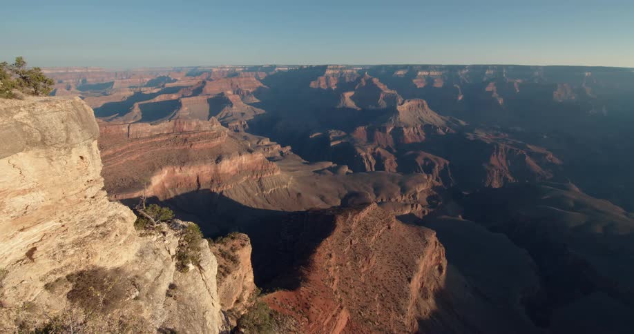 View from a ledge as the sun shines brightly upon rock formations and mountains under a clear sky