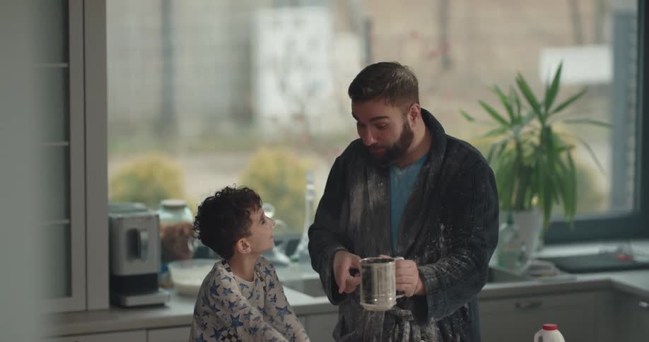 A boy and his dad have fun throwing flour at each other as they make breakfast