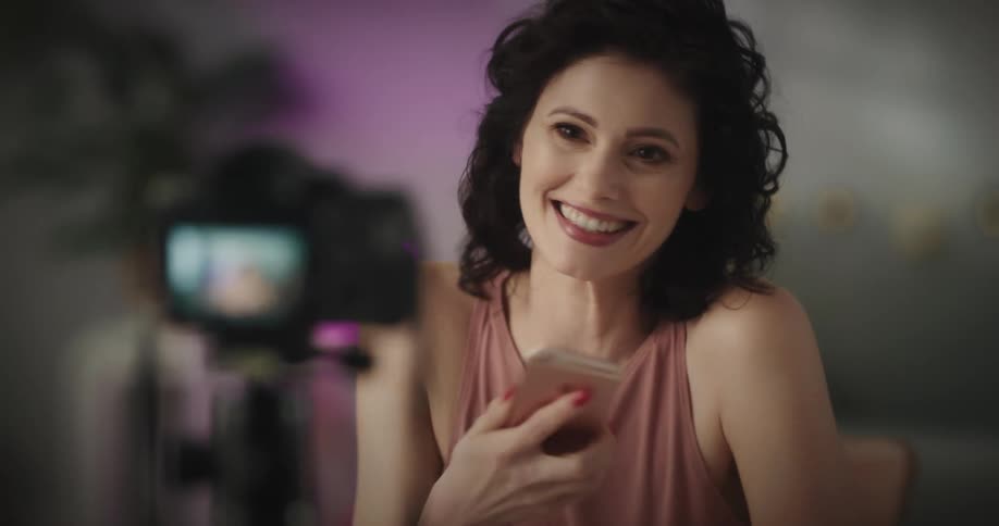A woman looks up from her smart phone and talks enthusiastically in front of a small digital camera