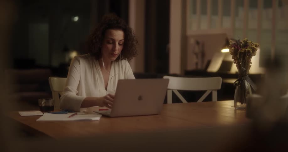 Woman with curly hair and white blouse sits and table working on laptop in the kitchen