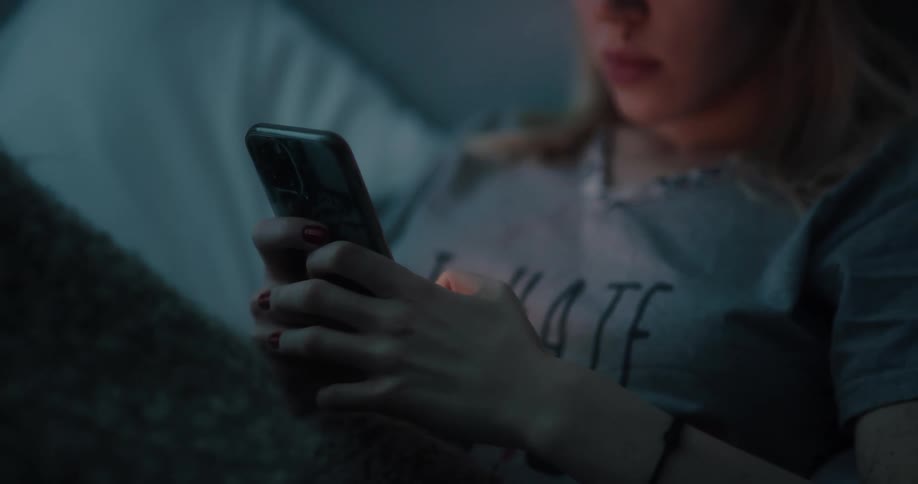 A young woman types on her smartphone while lying in bed in a dark room