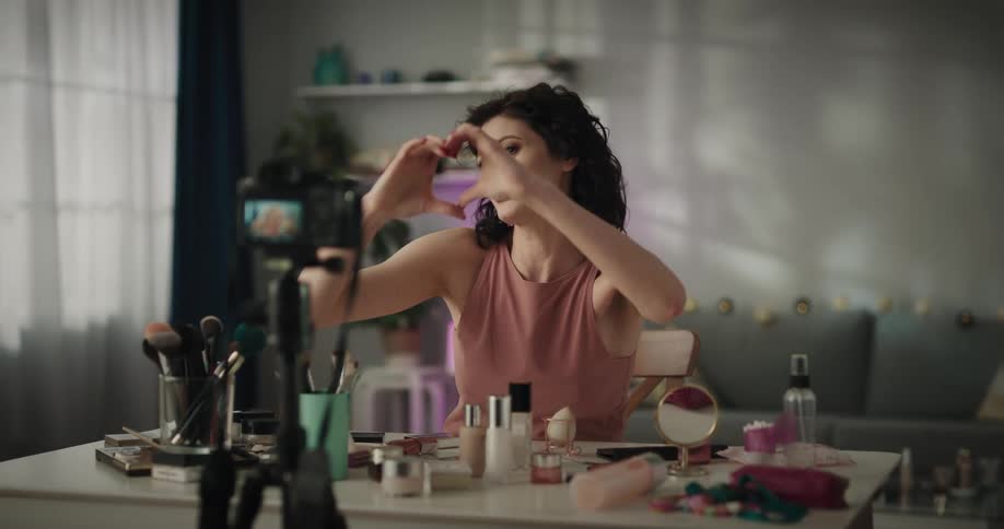 A woman sitting at a desk covered in makeup gestures for viewers to like and subscribe while recording herself with a camera