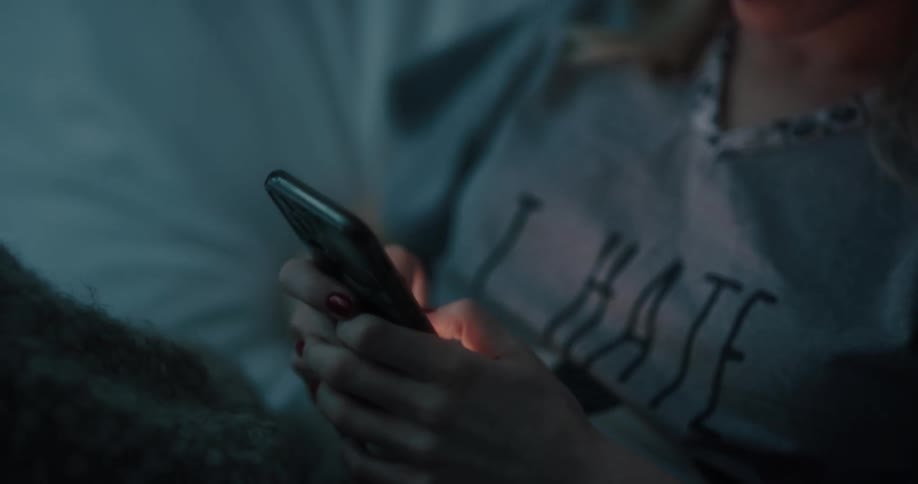 A woman is reclined in a bed while texting on a smartphone at night