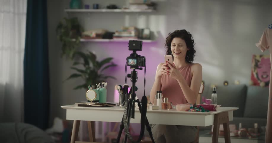 Social media influencer uses mobile phone at makeup table with tripod set to film