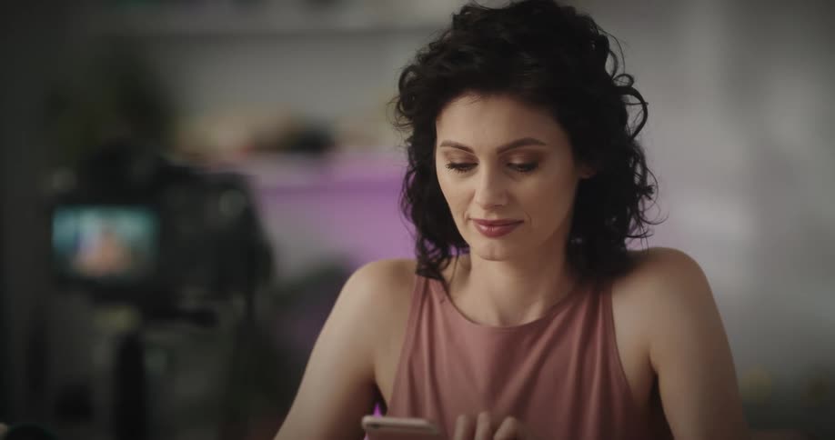 Camera video tapes woman sitting at table strewn with makeup as she smiles and texts