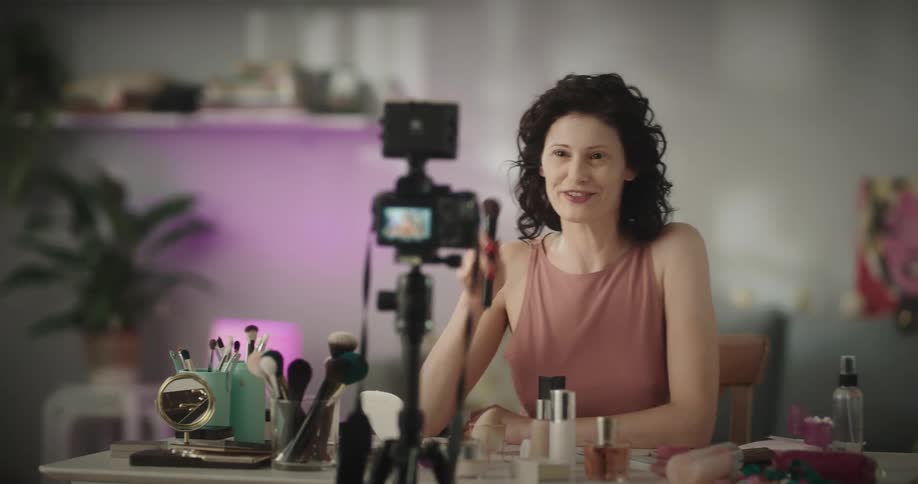 White woman sitting at a table full of makeups tools talking to a camera recording her