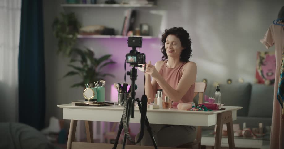 A woman is sitting at a table in a home while recording herself applying makeup and giving instructions in the daytime