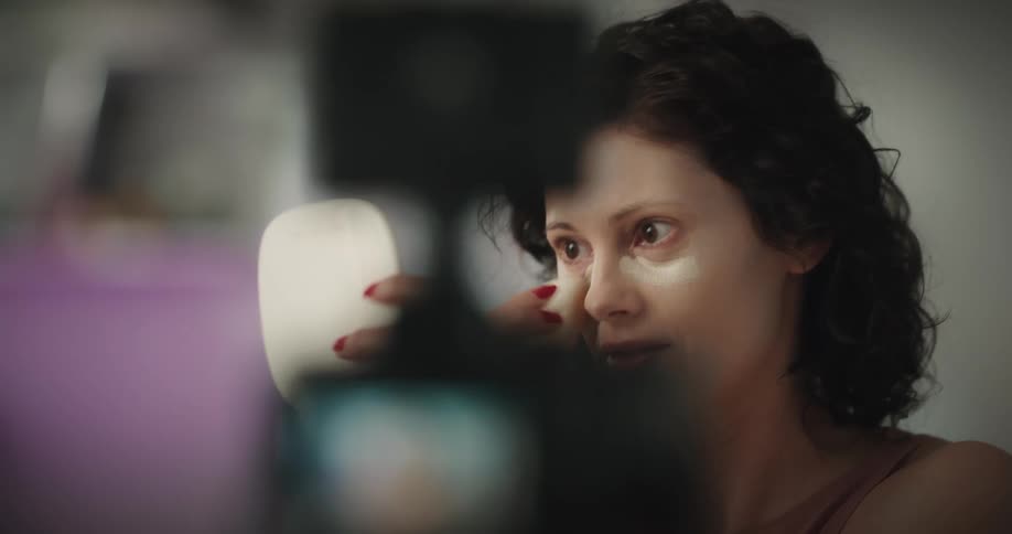 White woman holding a hand mirror and applying makeup while recording herself in a digital camera