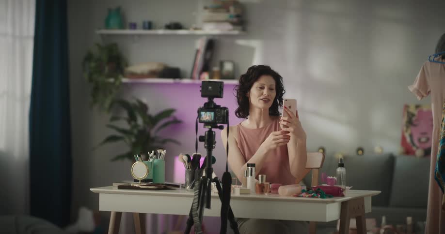 A woman films herself sitting at a makeup table holding up her smartphone