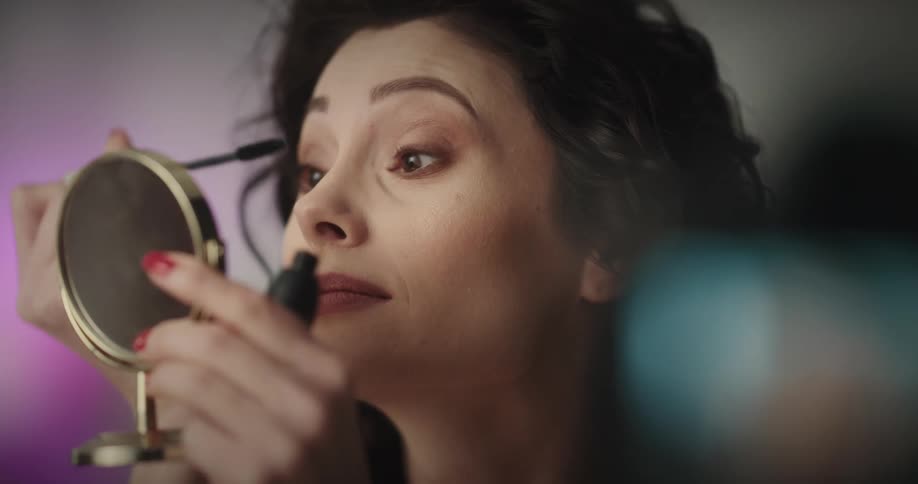 White woman holding a small mirror and applying mascara while she is recording herself in a digital camera