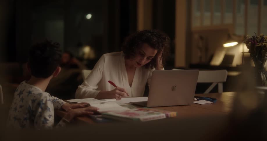 Mother and son sitting at the table while she is typing on the laptop and writing on a paper and he is reading a book
