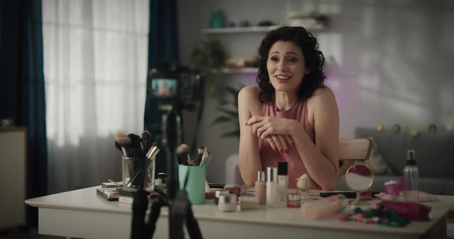 Woman sitting at table strewn with makeup gestures and talks into camera lens
