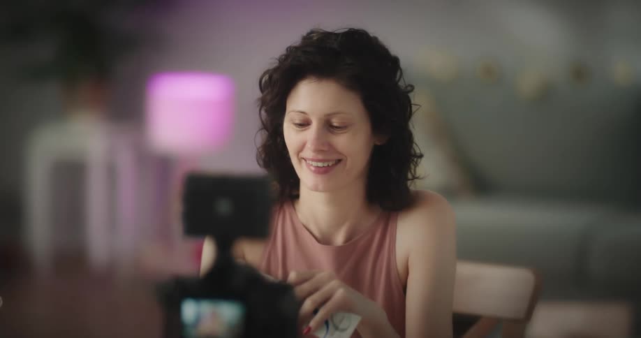 White woman sitting at the table full of makeup tools applying makeup while she is recording hersel in a digital camera