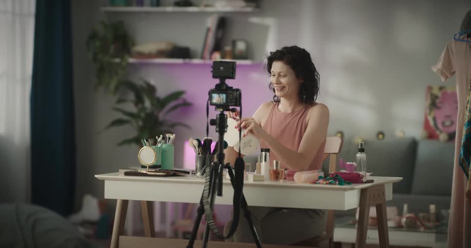 Woman sits at a table and demonstrates applying makeup while a camera on a tripod records her