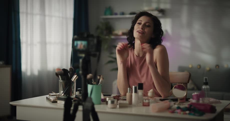 Female social media influencer turns on camera set on tripod and starts talking as she sits at makeup table