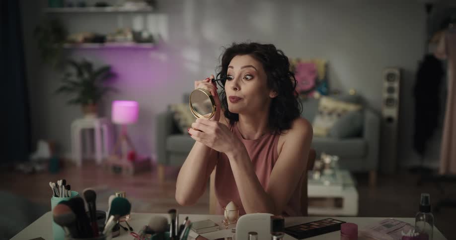 Woman talks as she applies mascara to her eye lashes while sitting at a table covered with makeup supplies