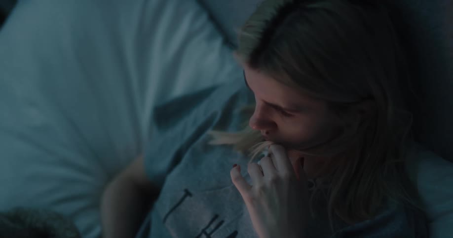 Blonde woman wearing gray t shirt uses smartphone in bed at night in bedroom