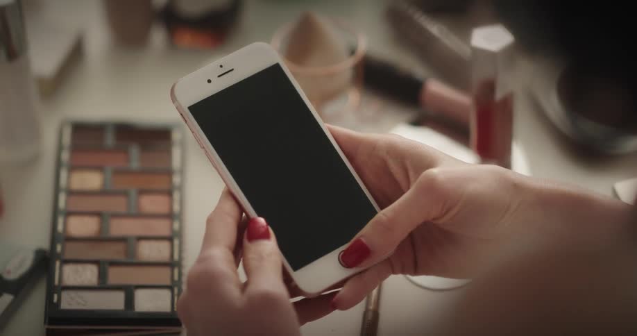 Womans hand holding a smartphone next to a table with a makeup palette and cosmetics