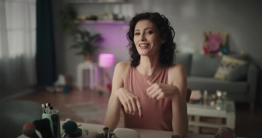 White woman sitting at a table full of makeup brushes and comestics applying mascara talking and gesticulating to the camera
