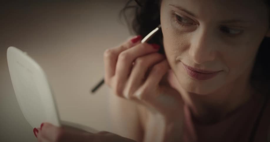 White woman holding a small hand mirror and applying makeup while talking and smiling