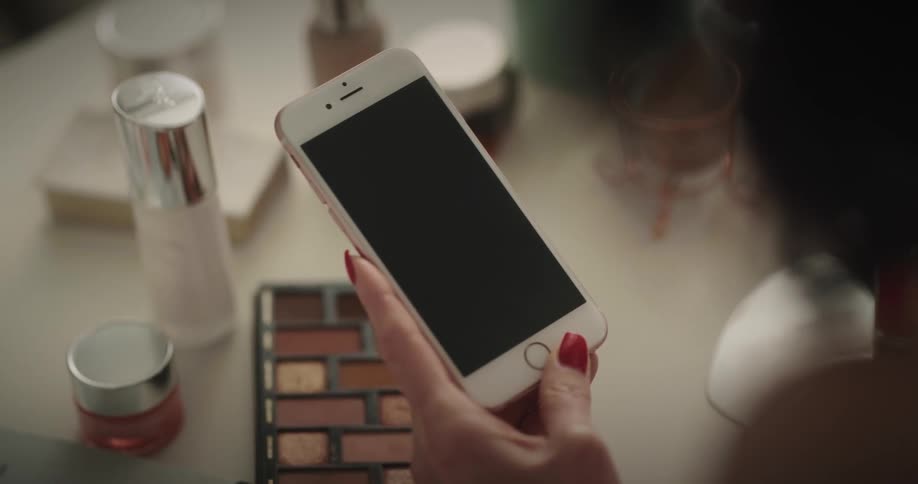 A woman holding a smartphone and touching the screen at her makeup table