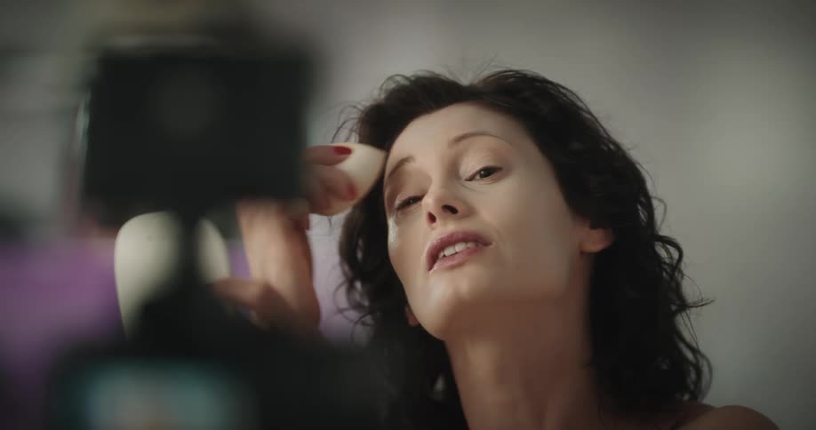 A woman holding up a mirror applying makeup with a sponge talking to an audience on camera