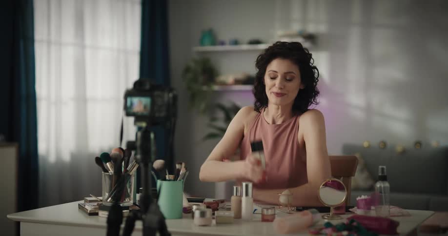 Woman sitting at a desk in her living room records a makeup tutorial while talking to the camera