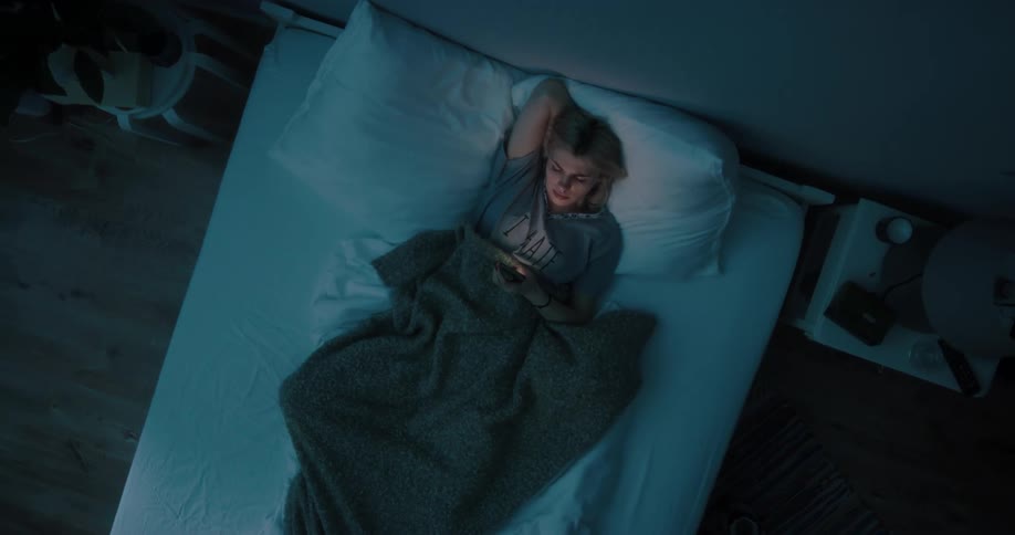 Blonde woman in gray t shirt lying in bed uses smartphone at night in bedroom