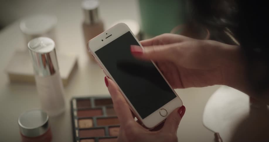 White womans hand using her smartphone in front of a table full of cosmetics and makeups