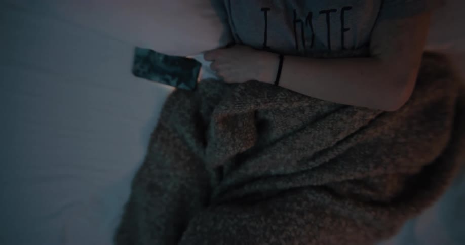 White woman lying in bed covering half of her body with a brown blanket using her smartphone