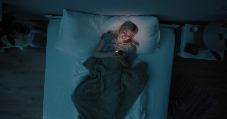 Blonde woman with gray t shirt lying in bed looks at ceiling and smiles using smartphone at night in bedroom