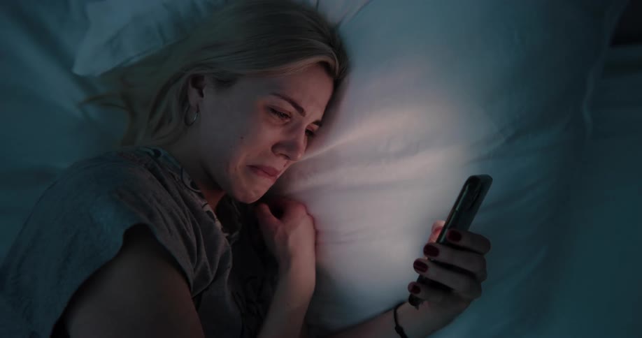 Blonde woman with gray t shirt looks at smartphone starting to cry and putting it under pillow at night in bedroom