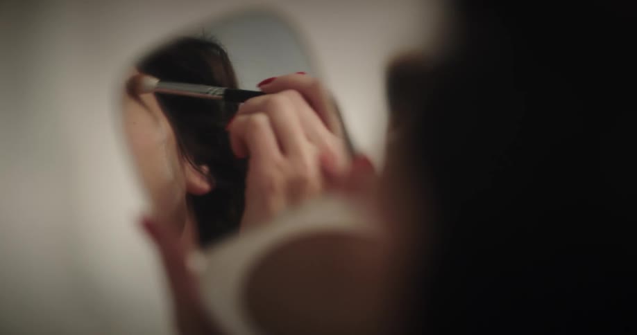 Woman with dark hair looks in a mirror as she applies eye shadow to her eyelids with a makeup brush