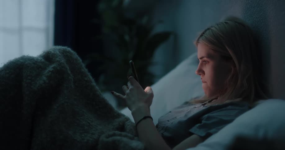 Blonde woman in gray t shirt sits in bed using smartphone at night in bedroom