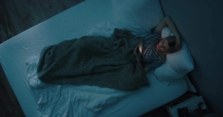 Blonde woman with gray t shirt lies in bed using smartphone at night in bedroom
