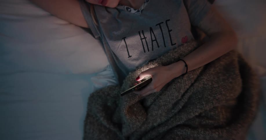 Blonde woman with gray t shirt lying in bed uses smartphone at night in bedroom