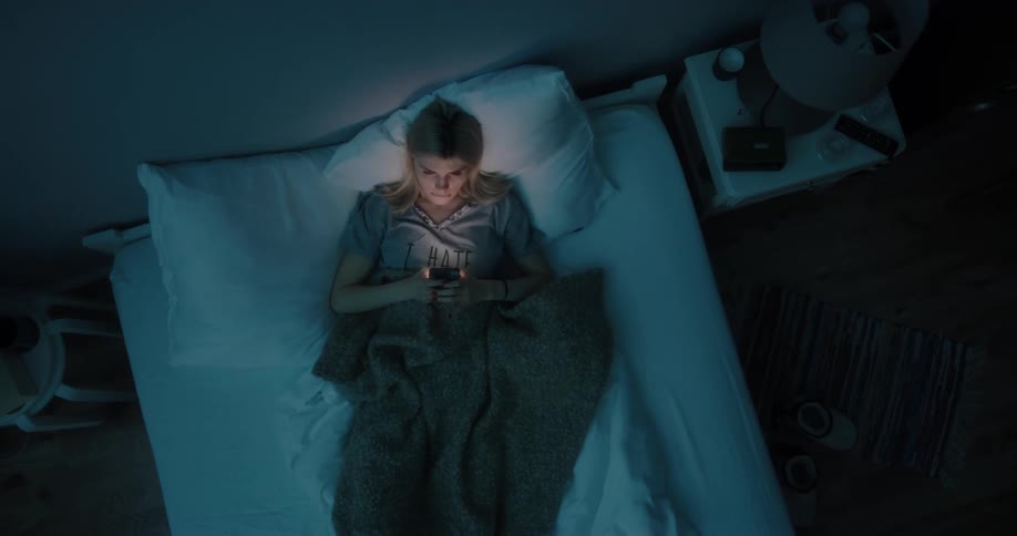 Overhead of young woman relaxing in bed texting on cell phone in the dark