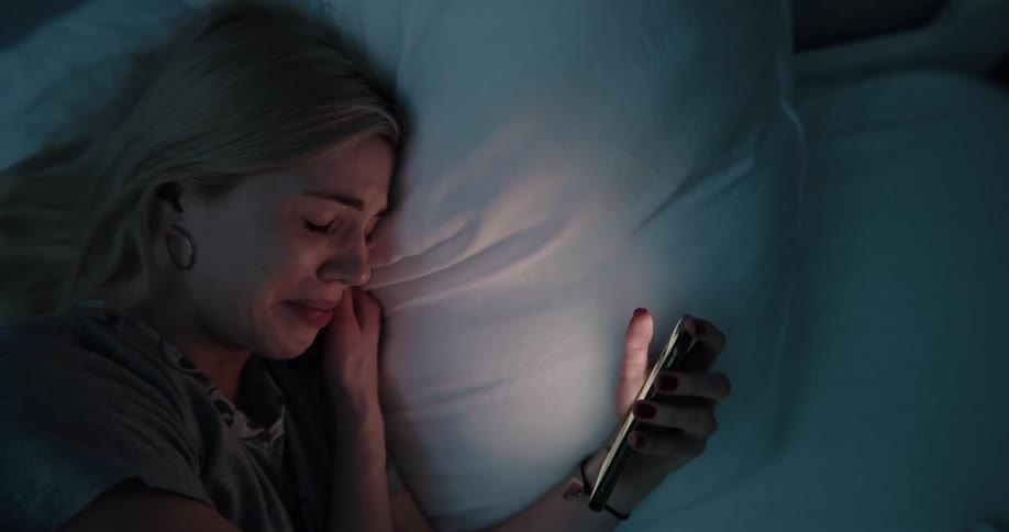 Blonde woman lying in bed uses smartphone and cries hard at night in bedroom