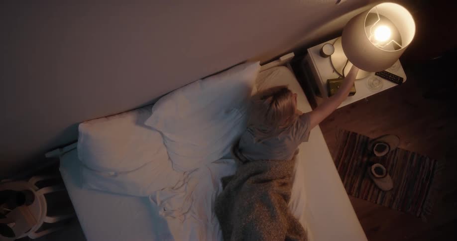 Blonde woman with gray t shirt lying in bed switches off bedside lamp at night in bedroom