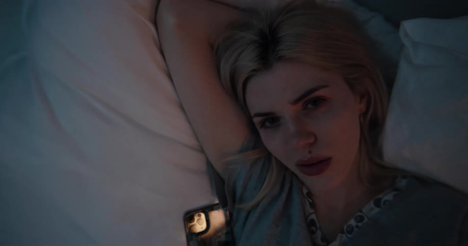 Blonde woman in gray t shirt lying in bed uses smartphone at night in bedroom