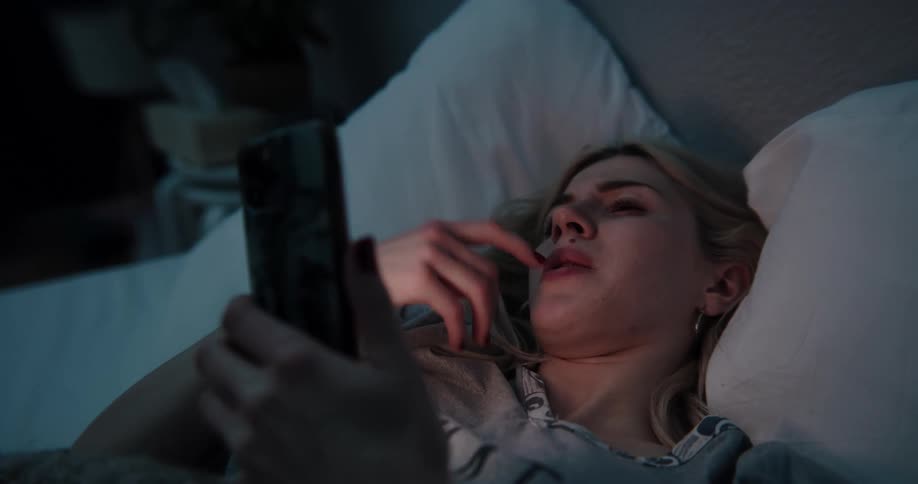 Blonde woman lying in bed cries and uses smartphone in bed at night