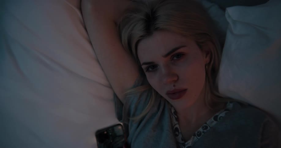 Blonde woman in gray t shirt lying in bed uses smartphone and looking at camera in bedroom at night