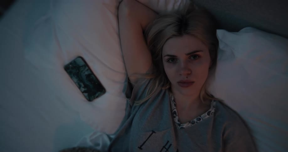 Blond teenaged girl lays on the pillows on a bed with her cell phone next to her