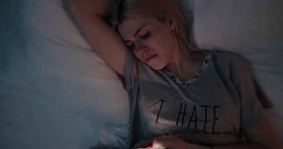 Young white woman lying in her bed scrolling through her smartphone with a sad looking face