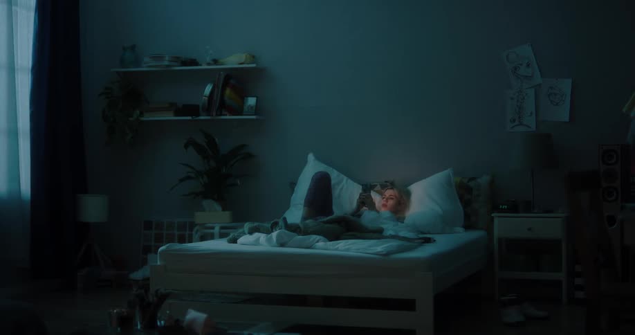 Blonde woman sitting in bed uses smartphone in bedroom at night
